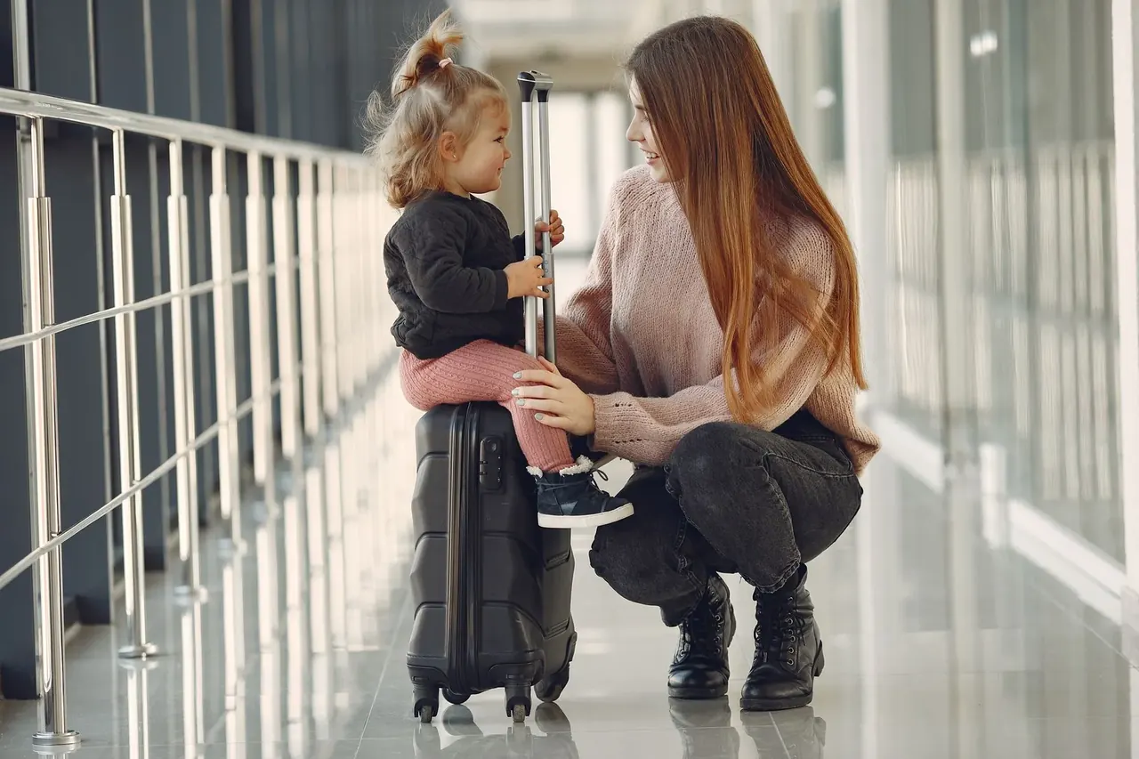 A mother and child pause beside their suitcase inside a bright airport terminal.